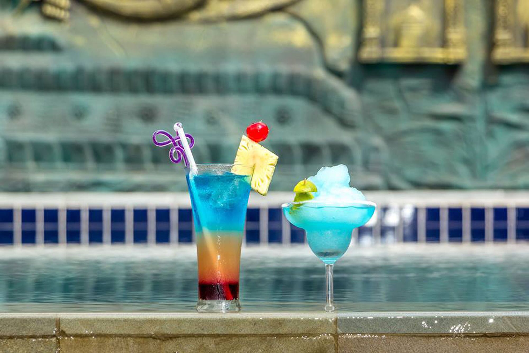Two cocktails on a poolside table with a fountain in the background.