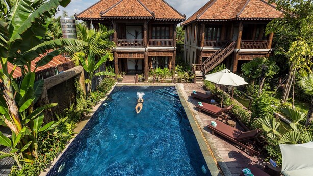 A pool area with a clear blue pool, wooden deck, lounge chairs, and two buildings with tiled roofs. - Java Wooden Villa by AIC (Siem Reap)