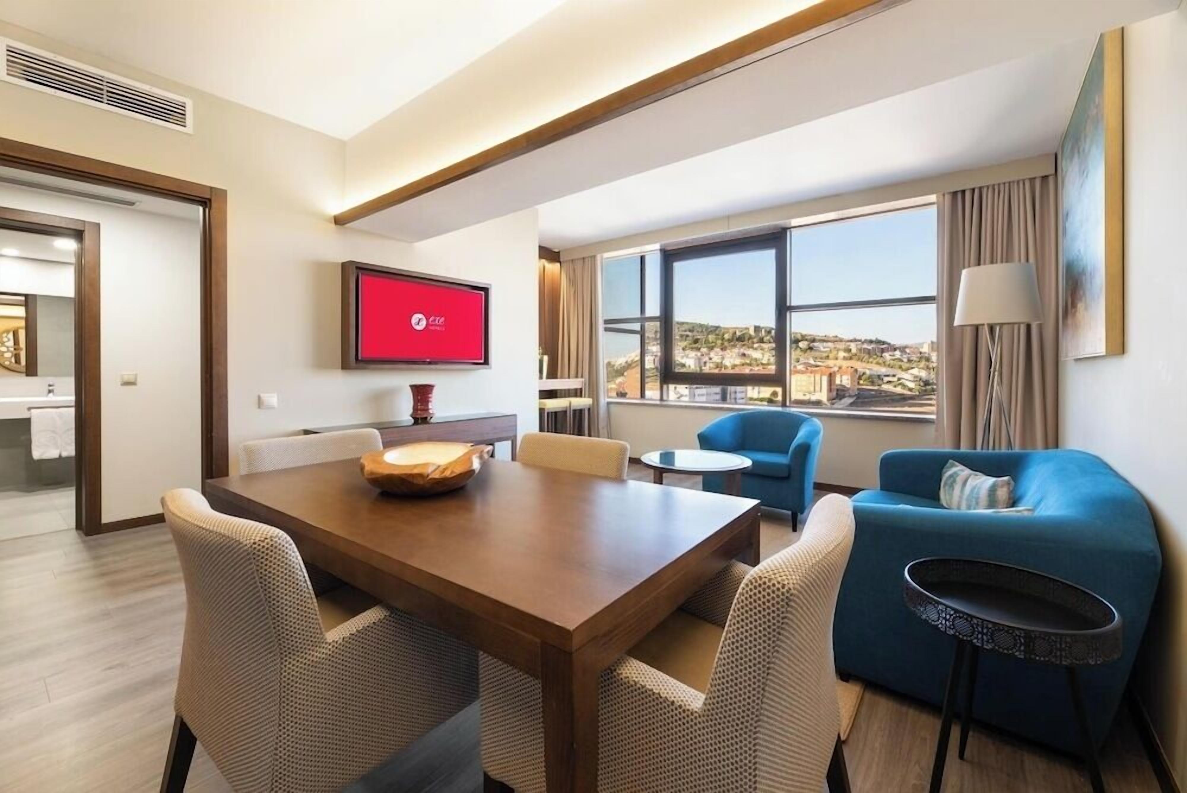 A hotel room with a wooden dining table, four chairs, a TV mounted on the wall, a sofa, a small round table, and a view of a cityscape through large windows.