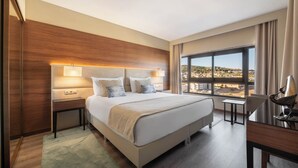 Une chambre d’hôtel moderne, dotée d’un grand lit, de tables de chevet, d’un bureau et offrant une vue sur le paysage urbain grâce à de grandes fenêtres.