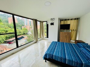 A bedroom with a large window, a bed with a patterned bedspread, a TV, and a wooden cabinet.