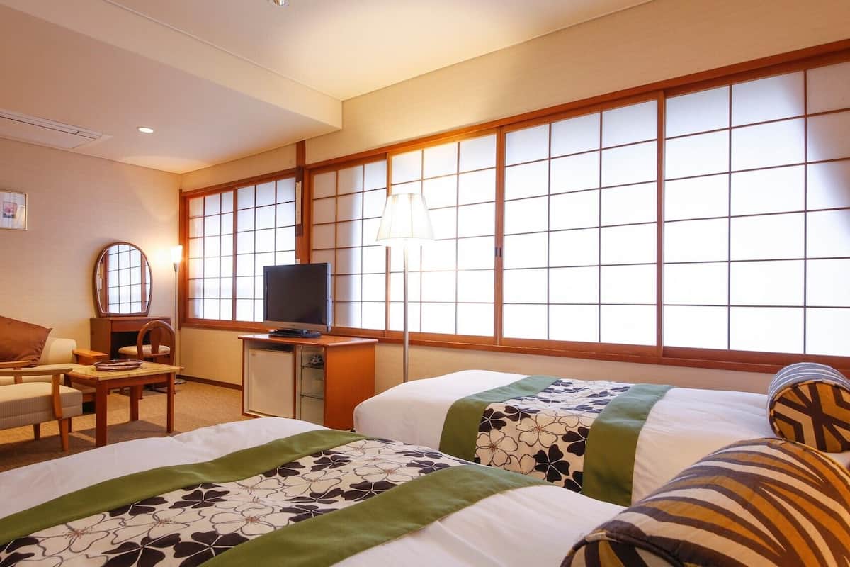 A hotel room with two beds, a desk with a TV, a chair, and a large window with a view.