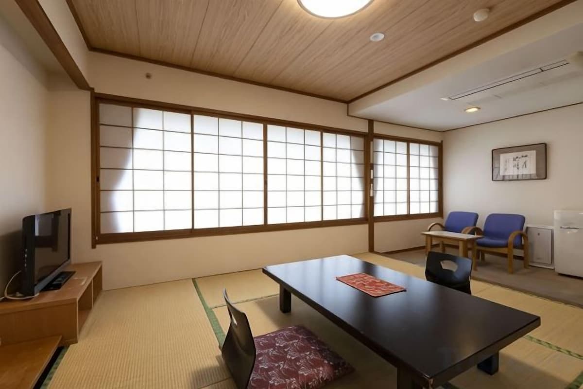 A traditional Japanese room with tatami flooring, a low table, two chairs, a television, and a large shoji screen.