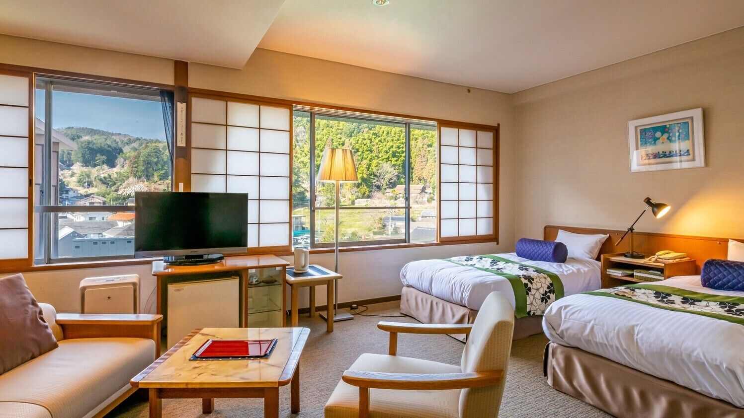 A hotel room with a television, two beds, a sofa, a desk, and large windows.