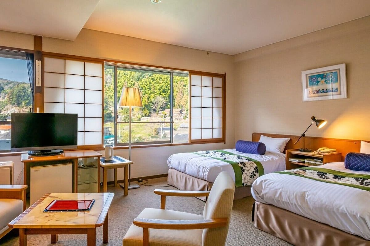 A hotel room with a television, two beds, a sofa, a desk, and large windows.