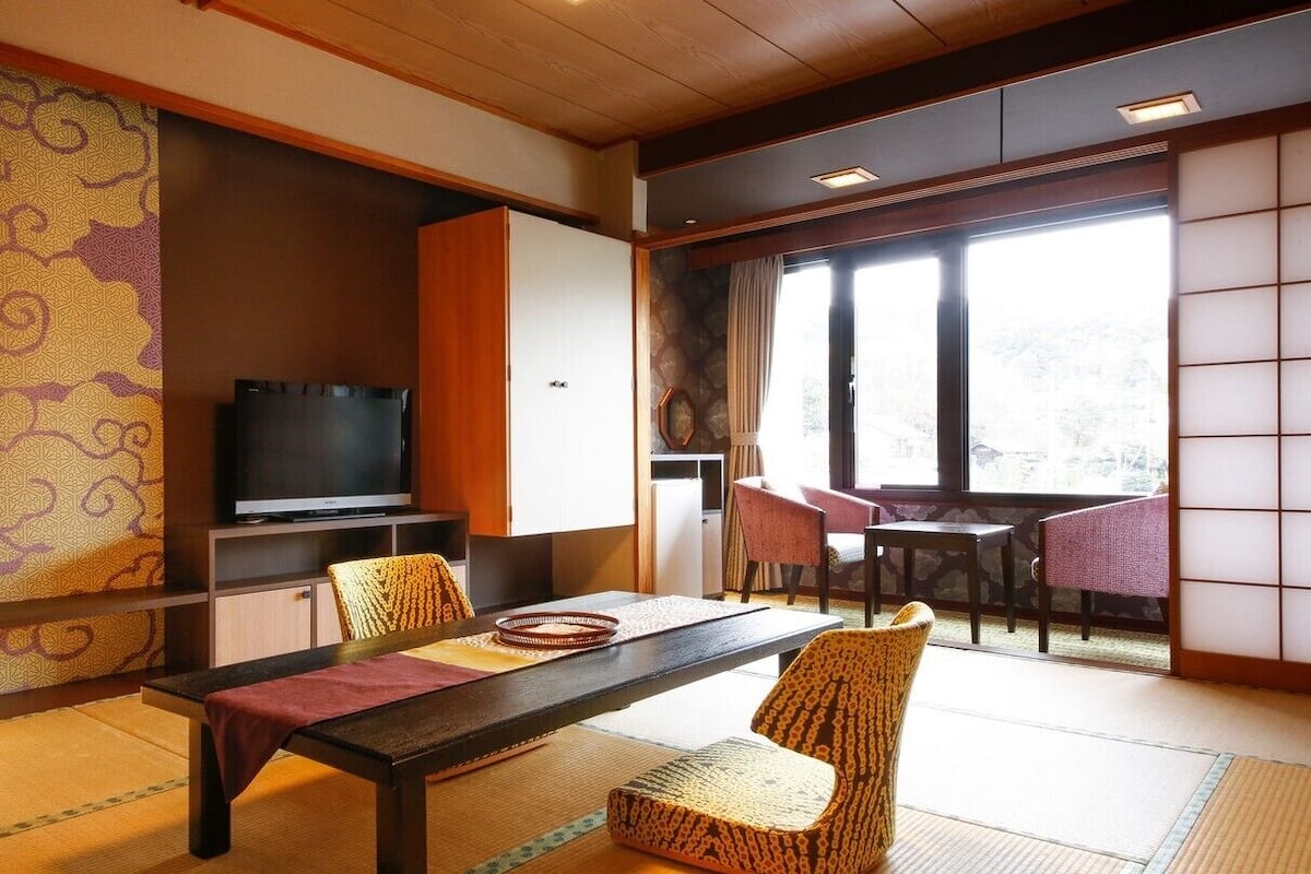 A traditional Japanese-style room with tatami flooring, a low table with cushions, a television, and a sliding door leading to a balcony.