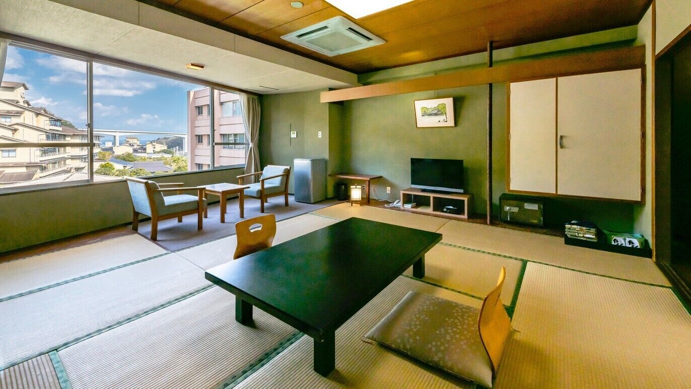 A room with tatami flooring, a low table, chairs, a television, and a view of buildings outside.
