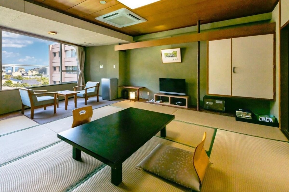 A room with tatami flooring, a low table, chairs, a television, and a view of buildings outside.