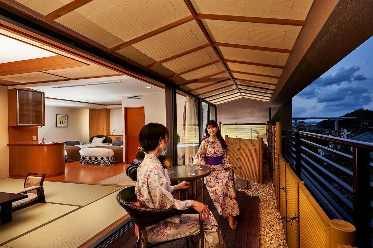 A room with a bed, a sitting area, and a balcony with a view of the city.