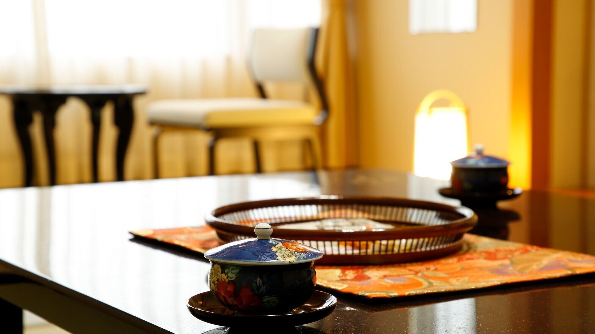 A traditional tea set on a table with a patterned mat, a chair, and a window in the background.