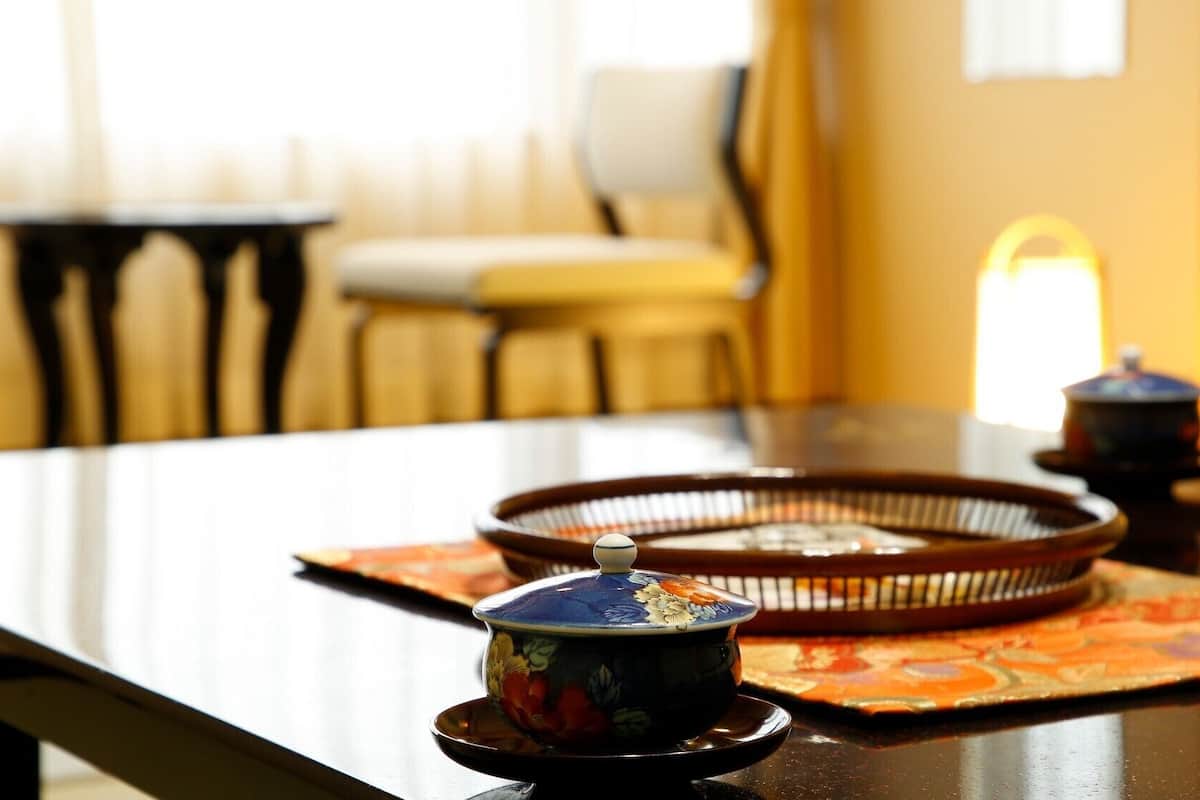 A traditional tea set on a table with a patterned mat, a chair, and a window in the background.