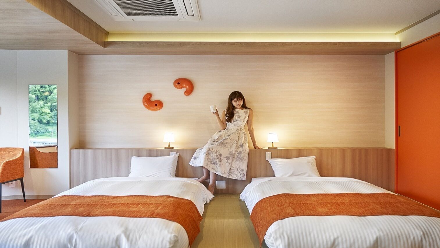 A hotel room with two beds, a wooden headboard, a mirror, a window with a view, and a wall with two orange shapes.