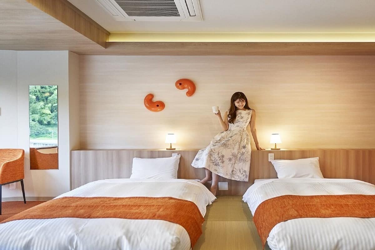 A hotel room with two beds, a wooden headboard, a mirror, a window with a view, and a wall with two orange shapes.