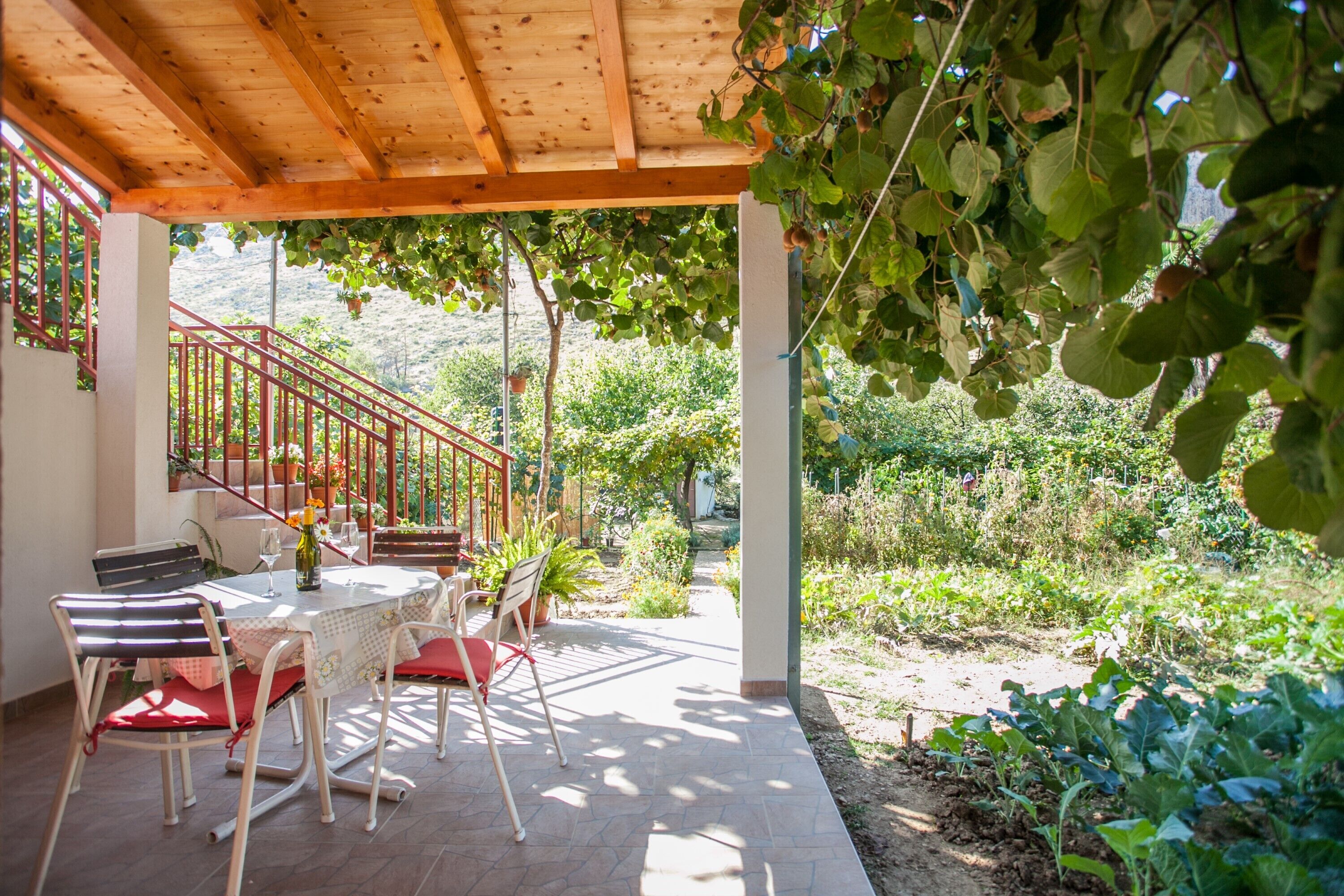 Um pátio externo com pérgola de madeira, uma mesa com cadeiras e vista para um jardim e colinas ao fundo.
