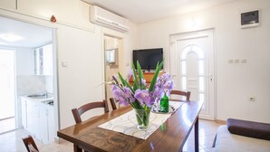 A dining area with a wooden table, chairs, and a vase of purple flowers. There is a kitchen visible in the background with a refrigerator and a television.