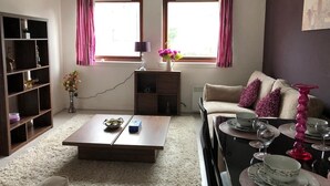 Ein Wohnzimmer mit einer Couch, Couchtisch und Esstisch, gedeckt mit Geschirr und Gläsern. Zwei Fenster mit Vorhängen, ein Bücherregal und eine Vase mit Blumen sind ebenfalls zu sehen.