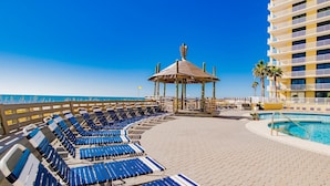 Uma área de piscina à beira-mar, com espreguiçadeiras, um gazebo e um edifício com varandas.