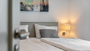 Ein ordentlich bezogenes Bett mit weißer und grauer Bettwäsche, eine Wandlampe sowie ein gerahmtes Bild darüber.