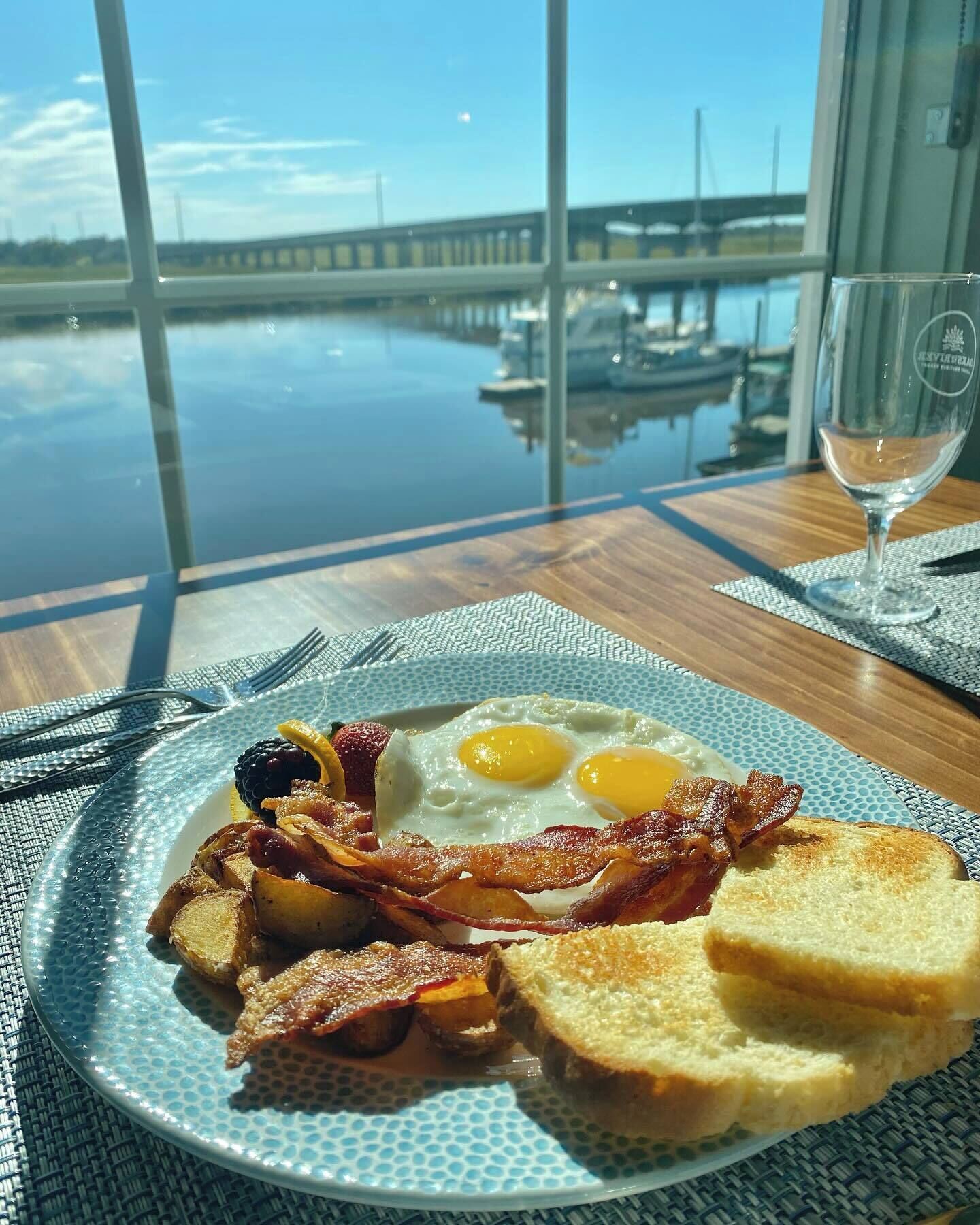 Ontbijtbord met eieren, spek, toast en fruit op een tafel met uitzicht op een jachthaven en een brug.