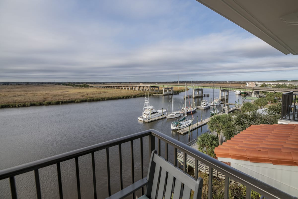 A view from a balcony overlooking a marina with boats docked, a bridge in the background, and a grassy area beside the water.