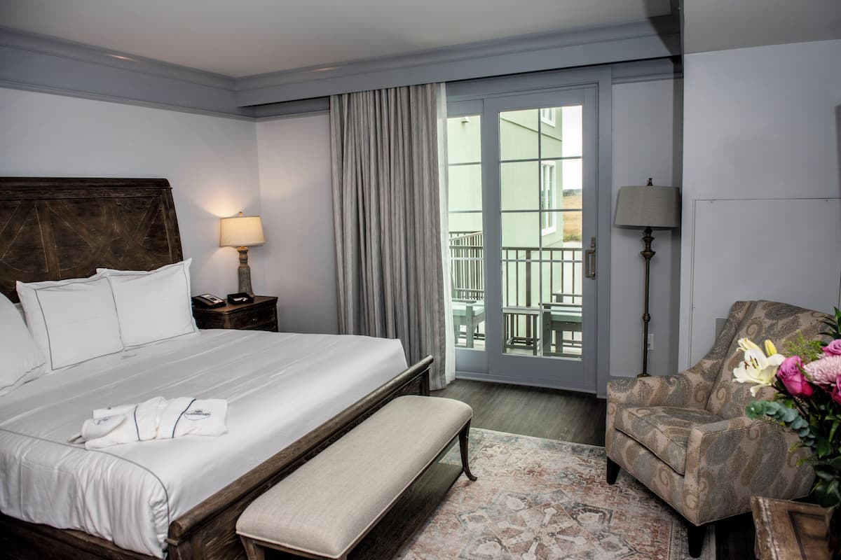 A hotel room with a large bed, a bench, a chair, a nightstand, and a view of the outdoors through a sliding door.