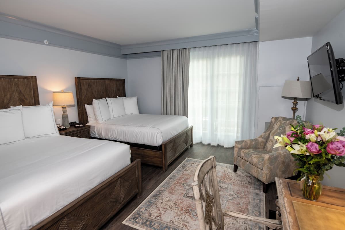 A hotel room with two beds, a wooden headboard, a wooden chair, a desk, a lamp, a TV, and a vase with flowers.