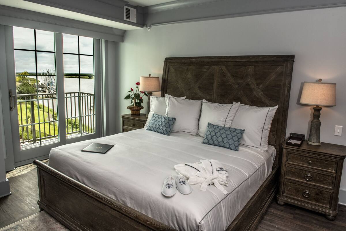 A large bed with a wooden headboard, white bedding, and a view of a balcony through a large window.