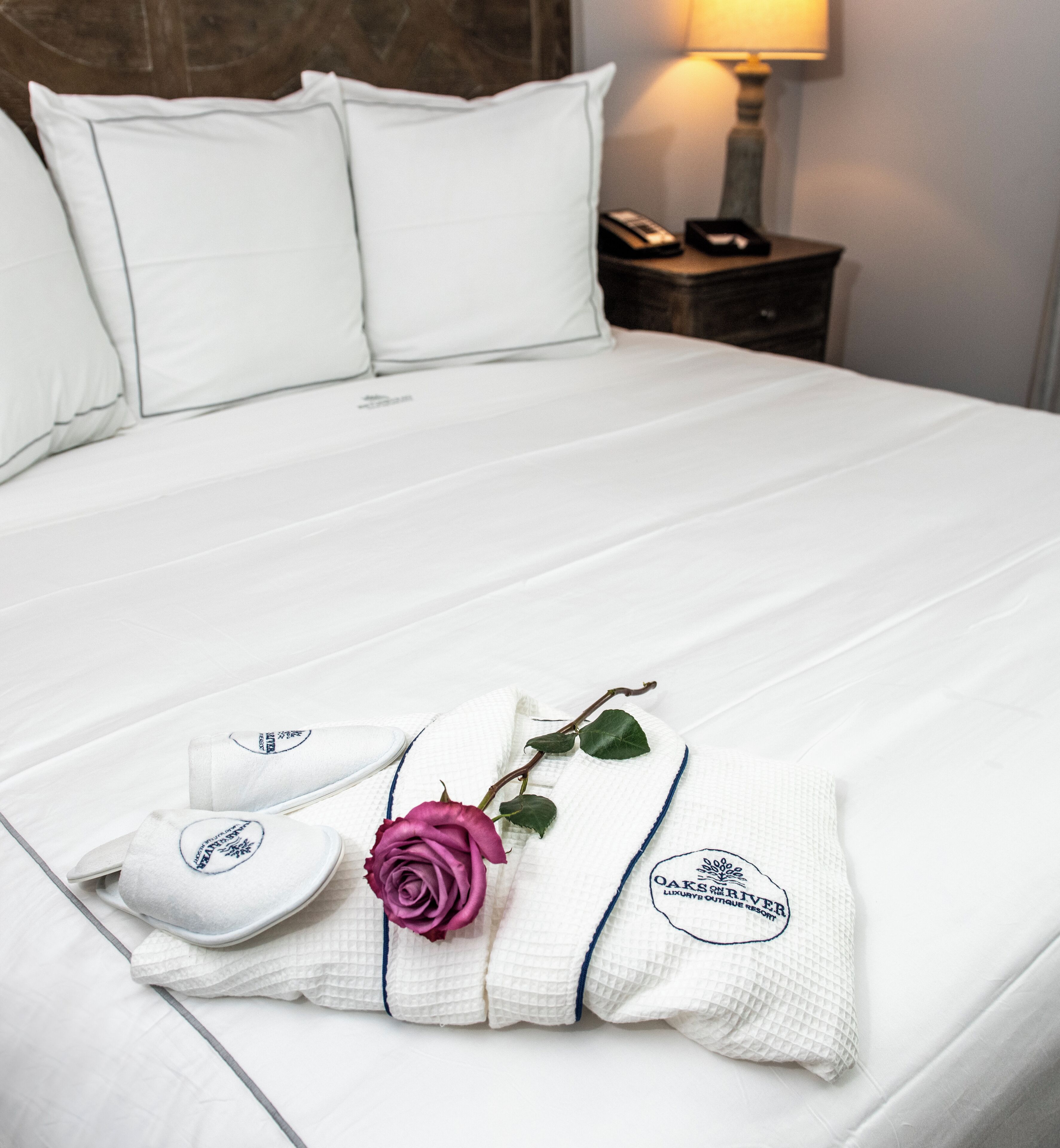 A neatly made bed with white linens, a decorative pillow with a logo, a folded towel, and a single rose.