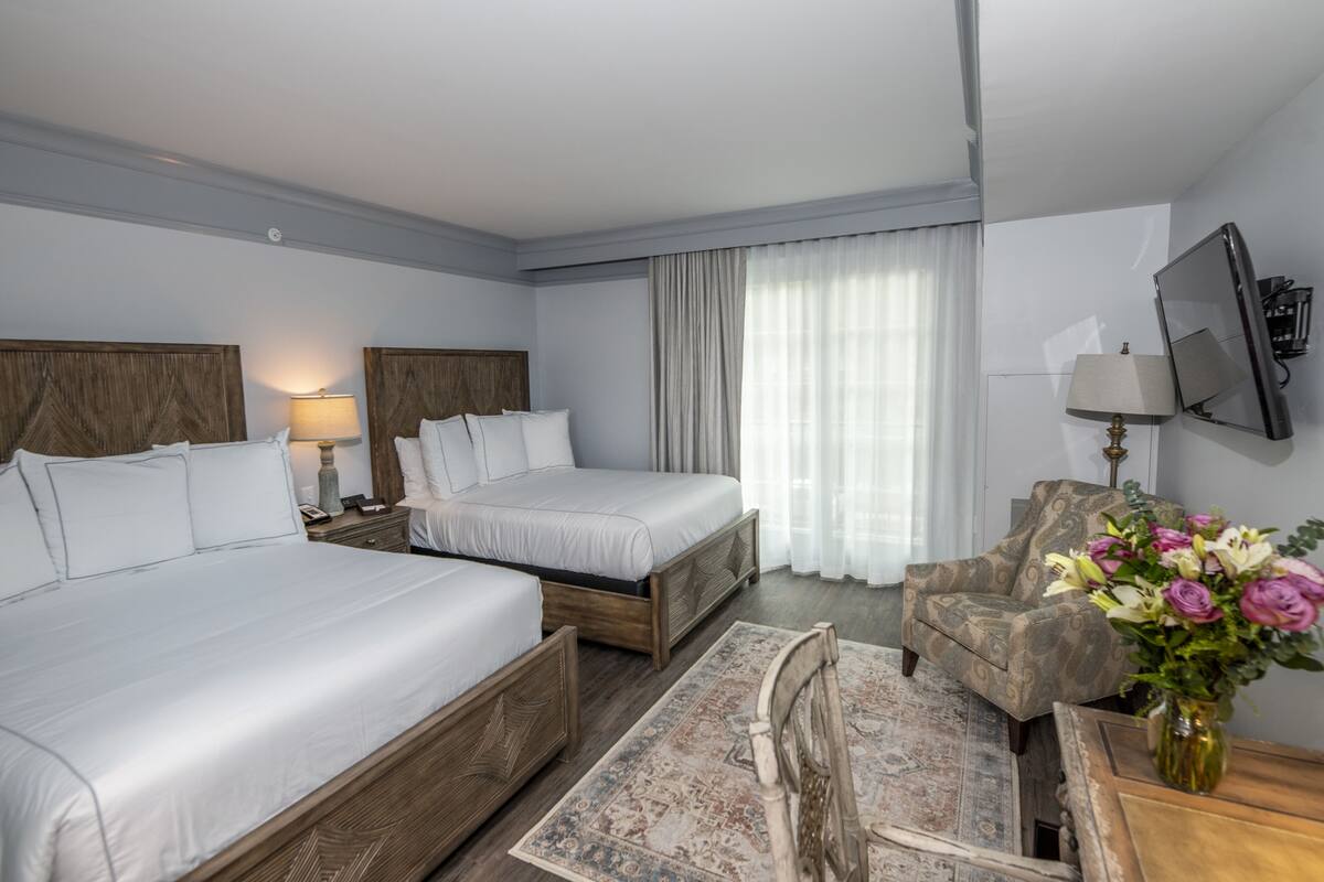 A hotel room with two beds, a TV, a chair, a desk, and a vase of flowers.
