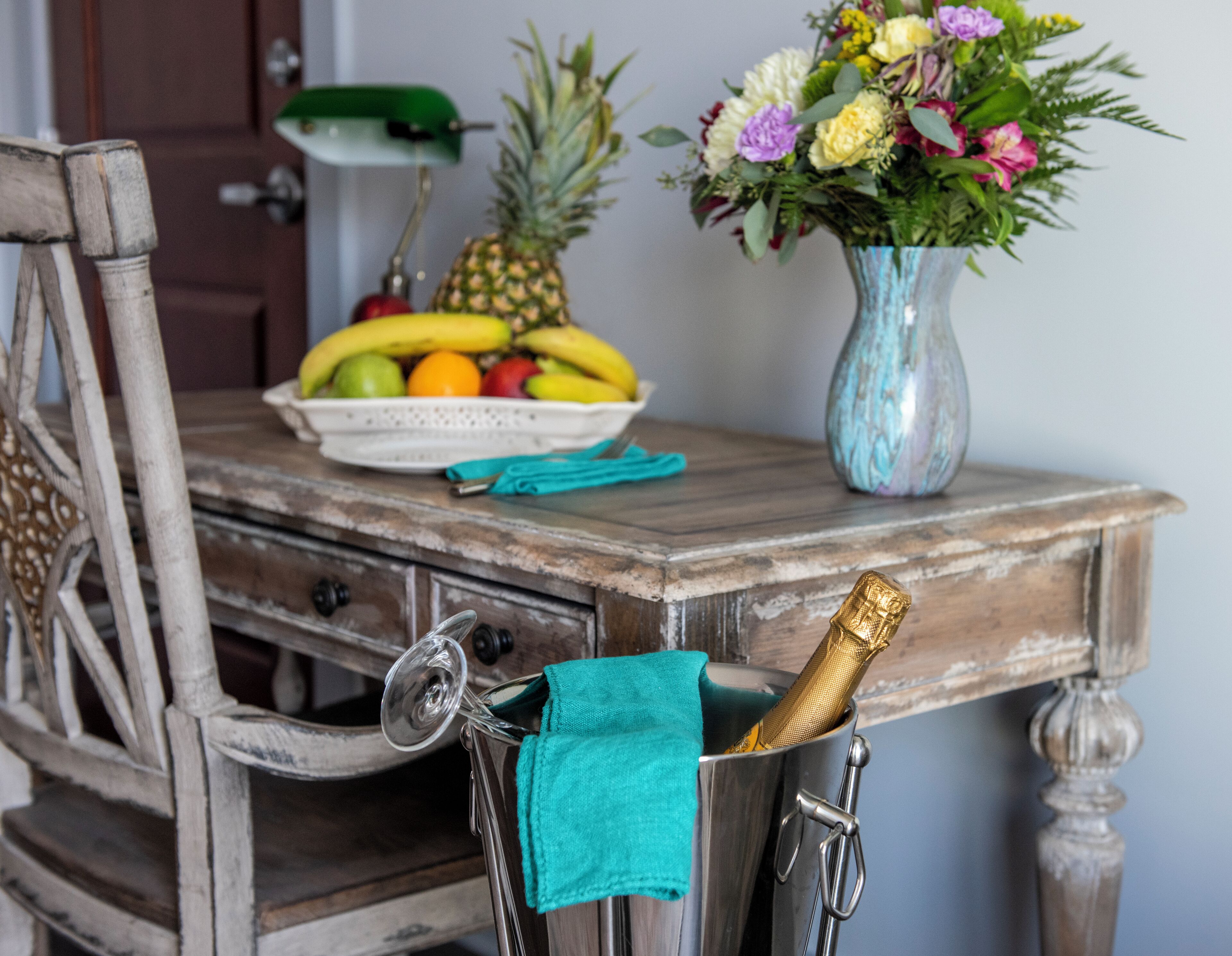 Un tavolo in legno rustico con una ciotola di frutta, un vaso di fiori e una bottiglia di champagne.