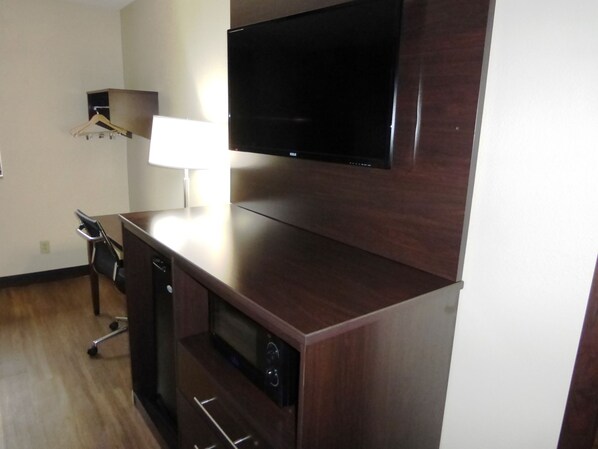 A modern living room with a flat-screen TV mounted on a wooden cabinet, a small table with a lamp, and a compact kitchenette with a microwave. - Red Roof Inn Bristol (Bristol)