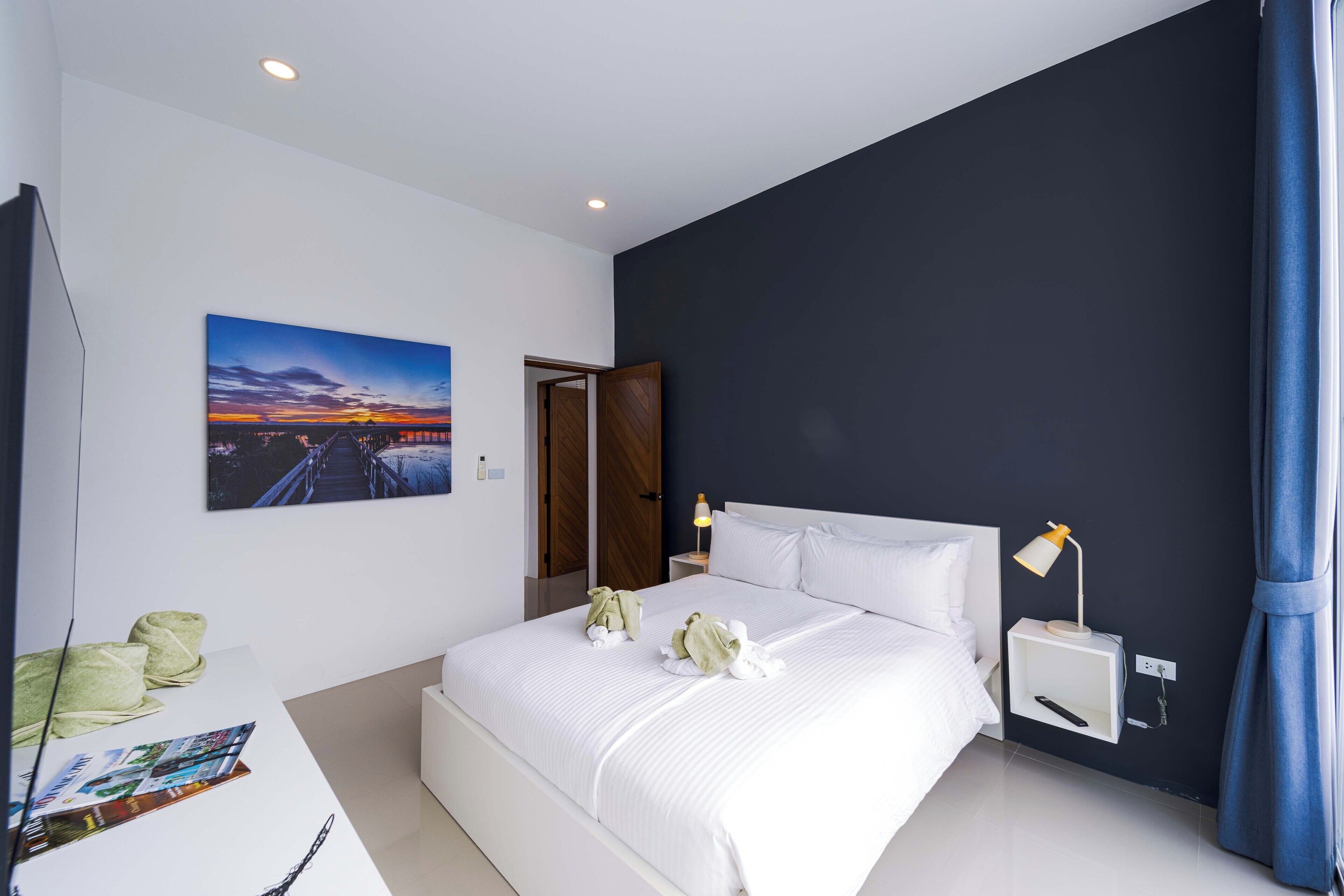 A modern hotel room with a large bed, a nightstand with a lamp, a painting of a sunset, and a desk with a magazine and a pen.
