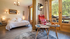 A bedroom with a bed, bedside lamps, a reading table with books and a plate of food, a red chair, and a large window with a view of trees. - Résidence le Chardonnet G 1BR for 4 w/Shared Pool & panoramic views, Argentière (Chamonix), France (Argentière (Chamonix))