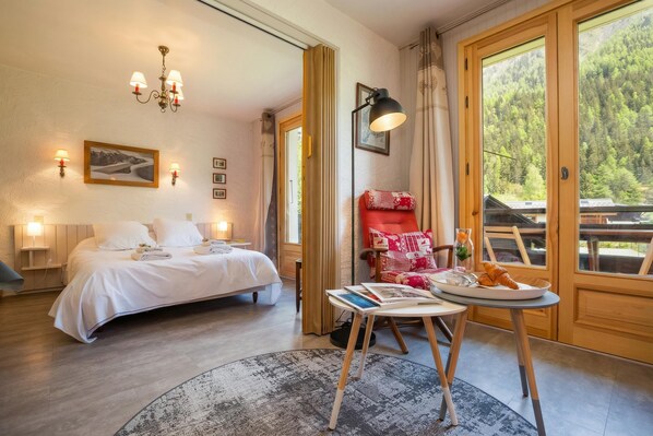 Ein Schlafzimmer mit Bett, Nachttischlampen, einem Lesetisch mit Büchern und einem Teller Essen, einem roten Sessel und einem großen Fenster mit Blick auf Bäume.