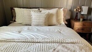 A neatly made bed with a white comforter and pillows, a wooden nightstand with a lamp and a vase with dried flowers, and a wooden headboard. - Cozy cabin retreat in rustic setting. Private lake, bonfire under the stars! (Lake)