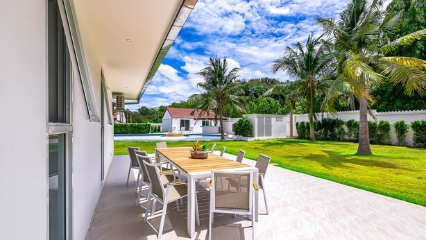 Une terrasse extérieure avec une table et des chaises en bois, une piscine en arrière-plan et une pelouse bien entretenue agrémentée de palmiers.