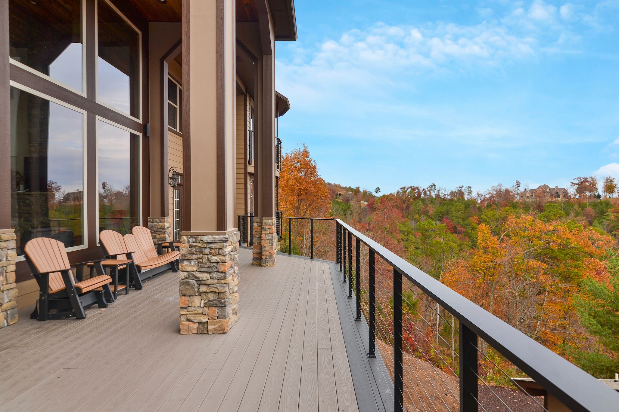 A wooden deck with Adirondack chairs, stone pillars, and a black railing overlooking a forest with autumn foliage.