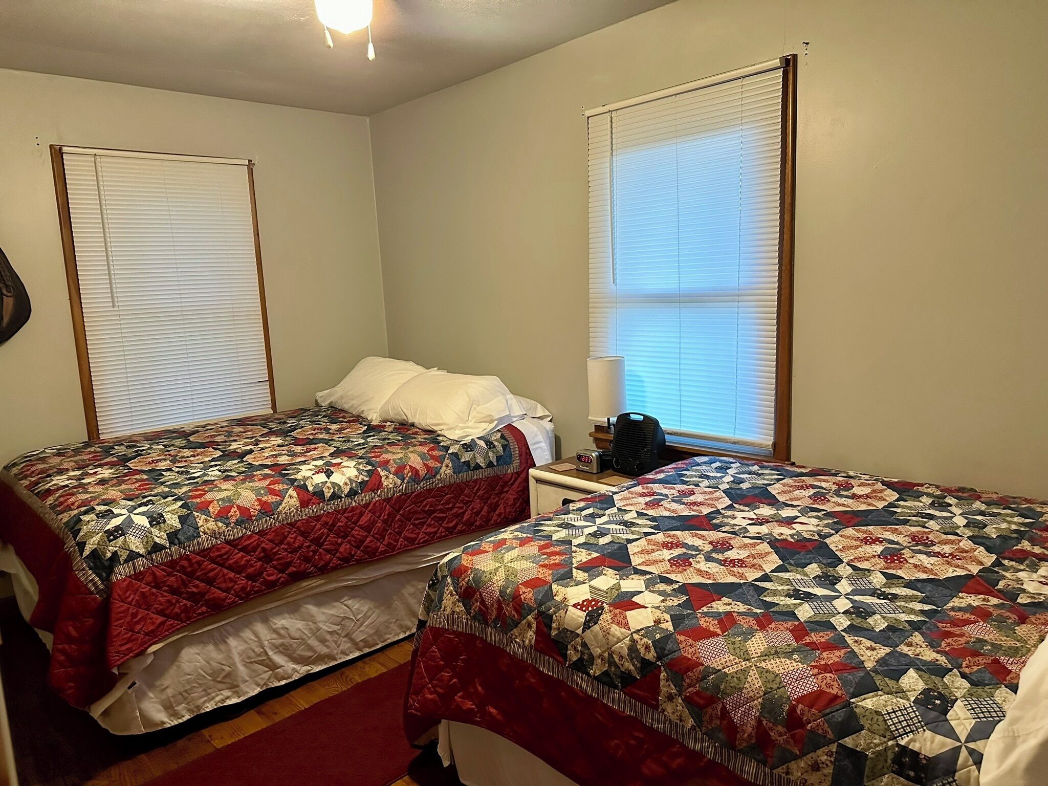 A room with two beds, each with a quilted bedspread, a bedside table with a lamp, and a clock.
