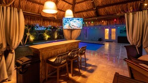 Un espace bar avec un comptoir en bois, des tabourets de bar, un écran de télévision et une piscine en arrière-plan.