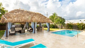 Un jardin avec une piscine, des chaises longues, un espace barbecue et un gazebo à toit de chaume.