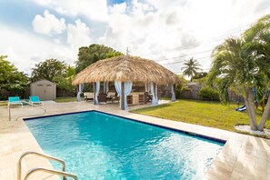 Una piscina con un pabellón de techo de paja, sillas de descanso y un cobertizo pequeño en un patio trasero.