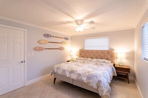 Un dormitorio con cama, ventilador de techo, ventana con persianas y elementos decorativos en la pared.