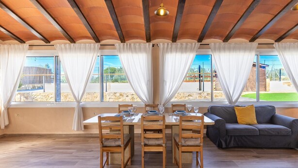 Un comedor con una mesa dispuesta para cuatro personas, sillas de madera, un sofá y vistas al exterior a través de grandes ventanales.