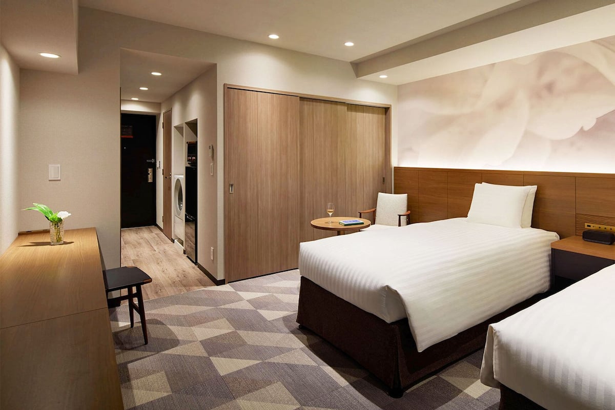 A hotel room with a large bed, a bedside table, a chair, a small table with a glass of wine, and a wall-mounted artwork.