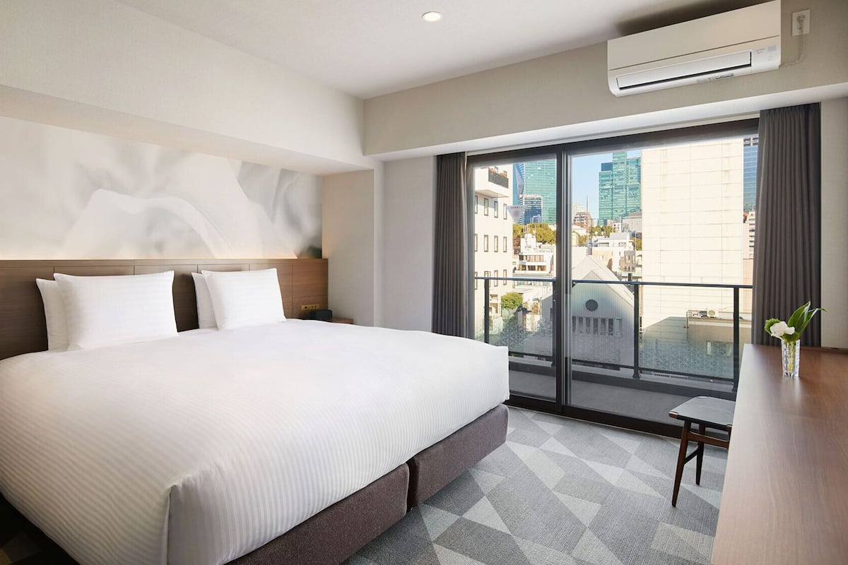 Ein Hotelzimmer mit einem großen Bett, einem Schreibtisch, einem Stuhl, einem Fernseher und einem Balkon mit Blick auf die Stadt.