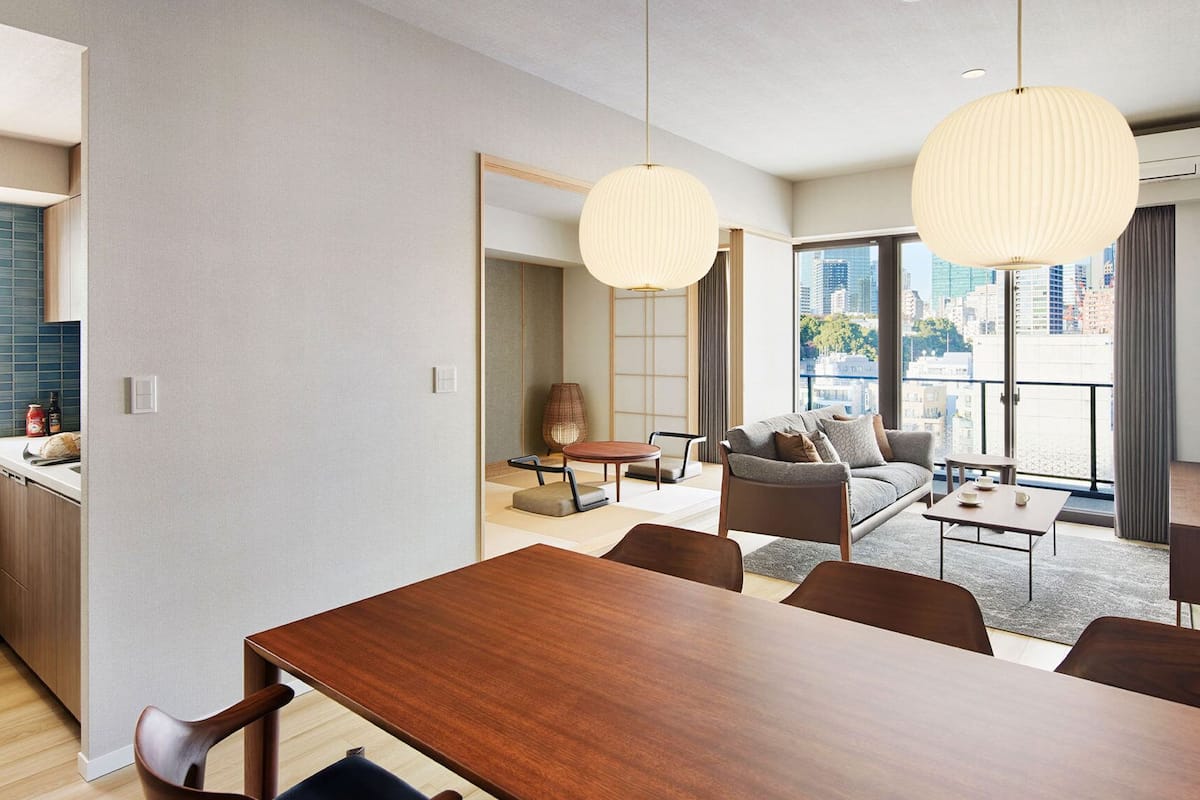 A modern living room with a sofa, coffee table, and dining table. There is a kitchen area with a counter and cabinets. The room has large windows with a city view.