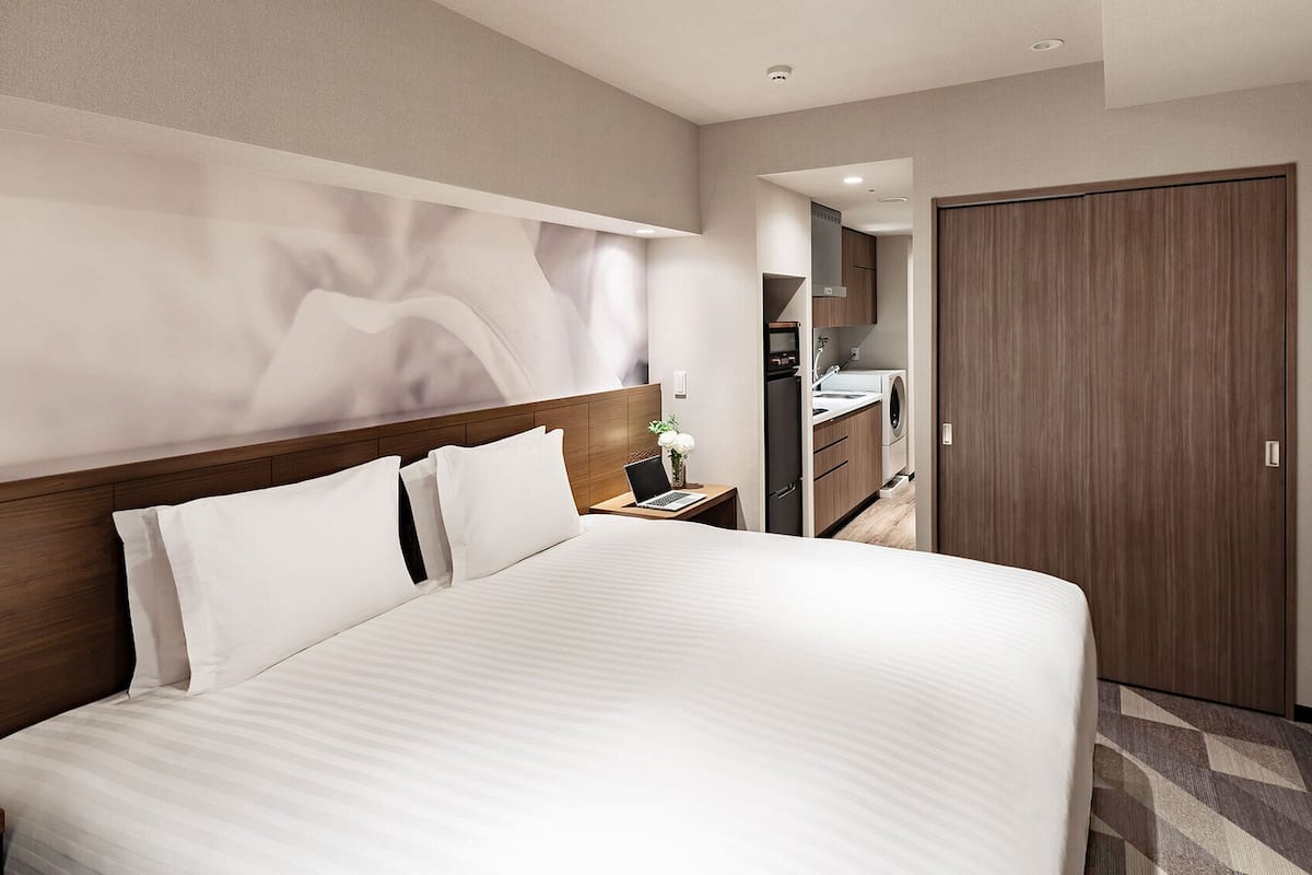 Ein Hotelzimmer mit einem großen Bett, einem Nachttisch, einem Schreibtisch mit einem Laptop, einem an der Wand montierten Fernseher und einem durch eine geöffnete Tür einsehbaren Badezimmer.