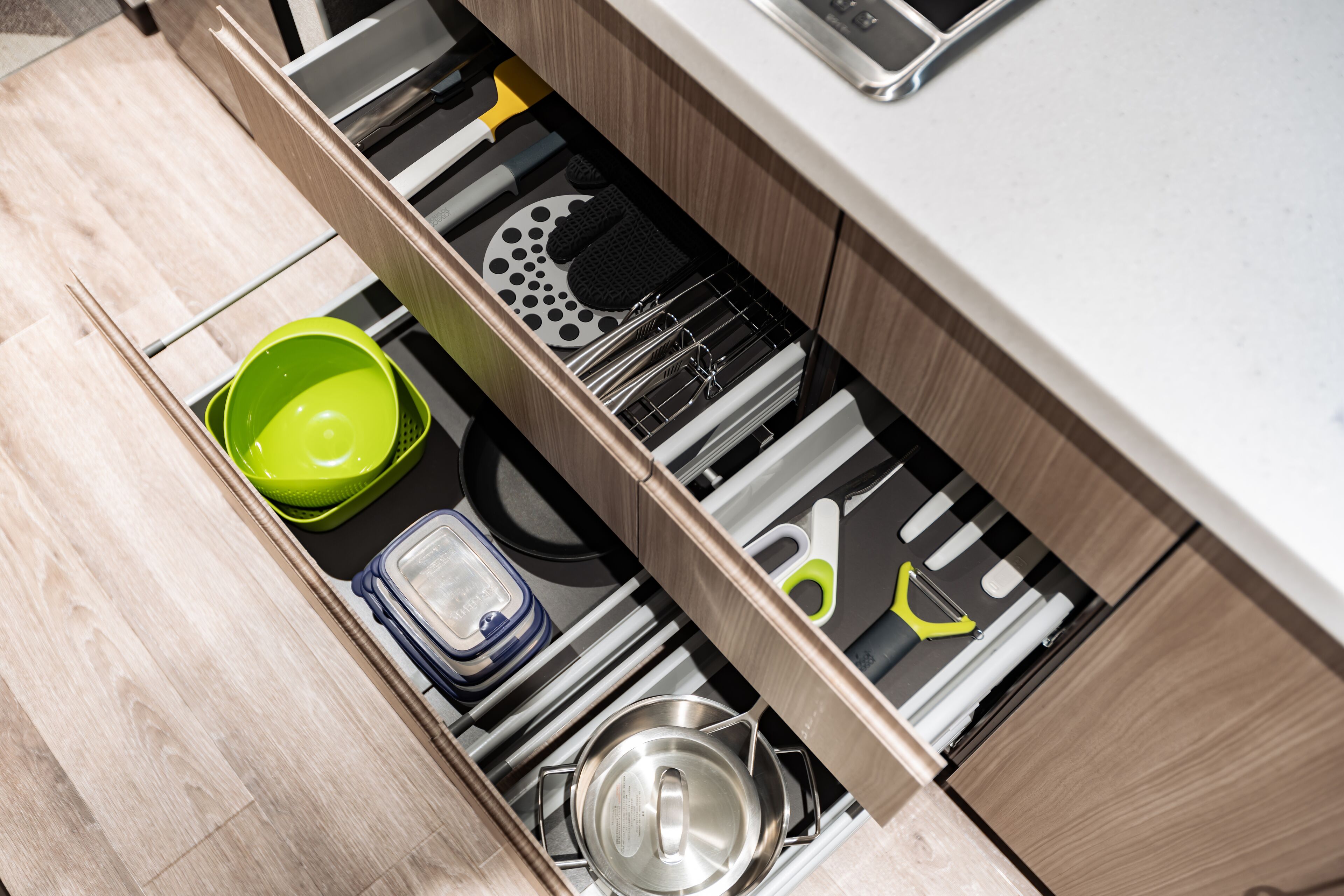 Un cajón de cocina con utensilios, un tazón verde y una olla de acero inoxidable.
