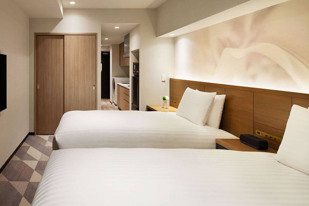 A hotel room with two beds, a TV, a wooden wardrobe, and a bathroom visible in the background.