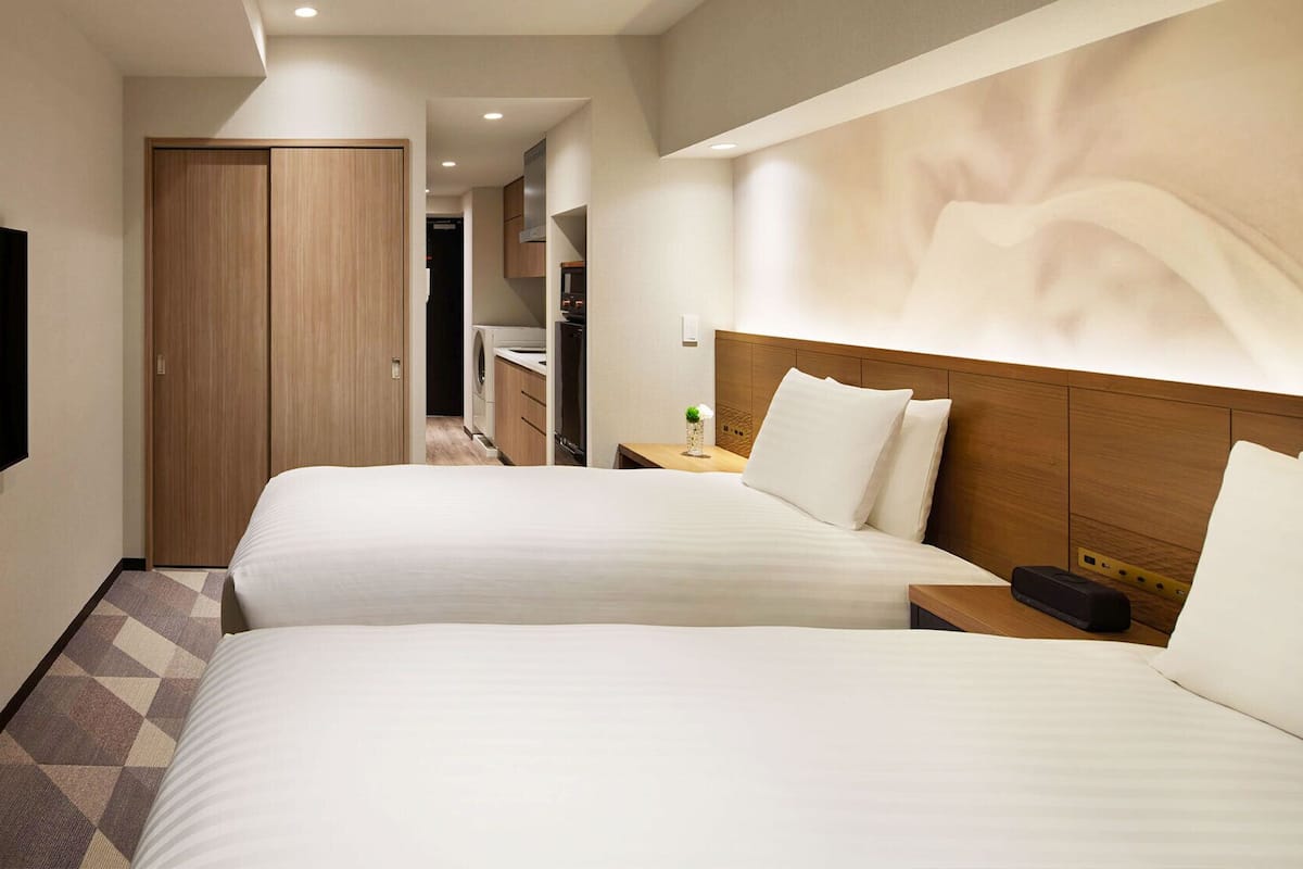 A hotel room with two beds, a TV, a wooden wardrobe, and a bathroom visible in the background.